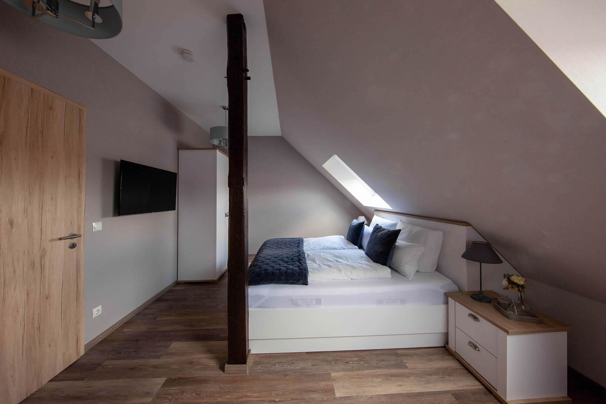 Ein Schlafzimmer mit schrägverdeckter Decke, einem Bett mit weißer Bettwäsche, einem Nachttisch und einem Holzschrank.