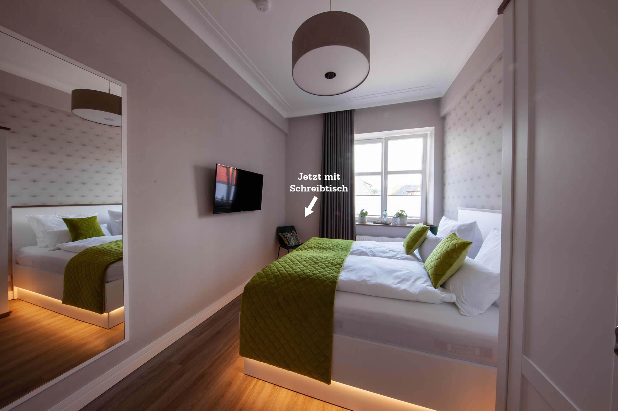 Ein modernes Hotelzimmer mit einem großen Spiegel, einem Flachbildfernseher, zwei Betten mit grün-weißer Bettwäsche, einem Fenster mit Vorhängen und einer Deckenleuchte.
