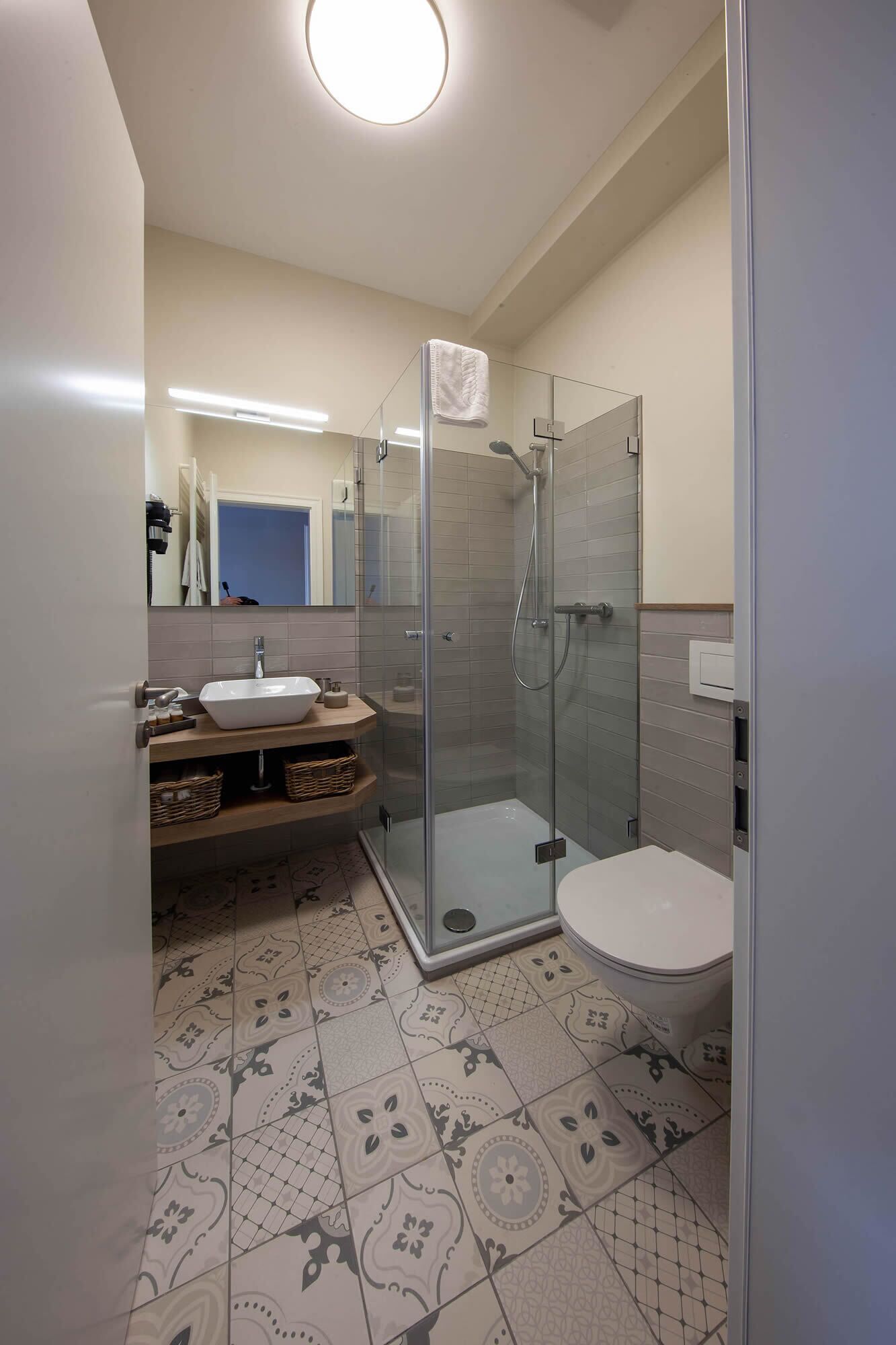 Un baño moderno con ducha envuelta en vidrio, lavamanos con espejo y baldosas de suelo estampadas.
