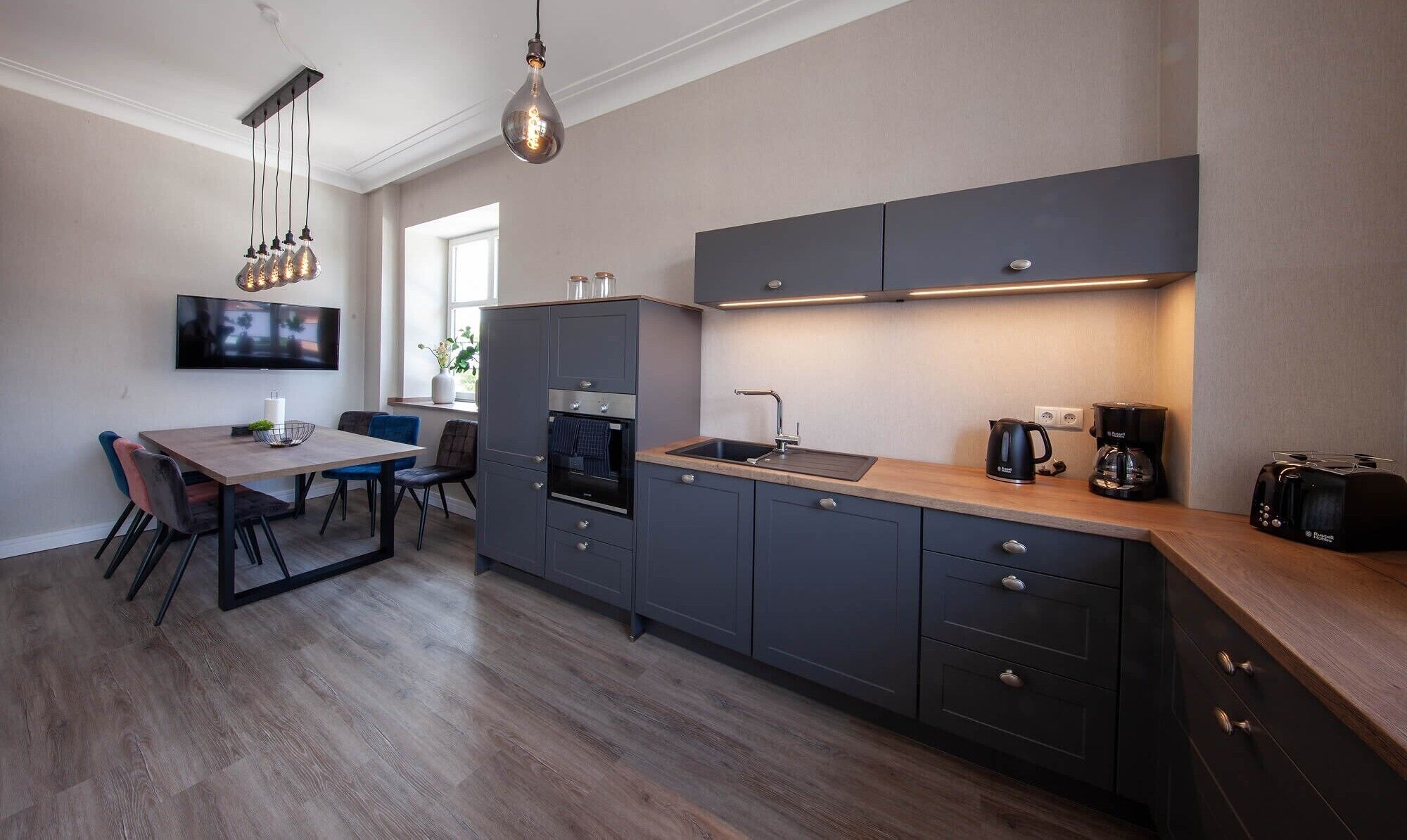 Een moderne keuken met grijze kasten, een houten aanrechtblad en een inbouwoven. Er is een zithoek met een tafel en stoelen, en een aan de muur bevestigde televisie.