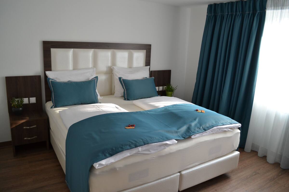 Una cama bien hecha con cubrecama color turquesa y almohadas blancas, cabecera de madera, mesita de noche con una planta y ventana con cortinas azules.