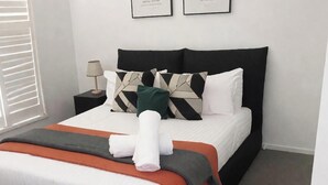Cama bem arrumada com travesseiros, abajur e toalha enrolada sobre um almofada laranja.