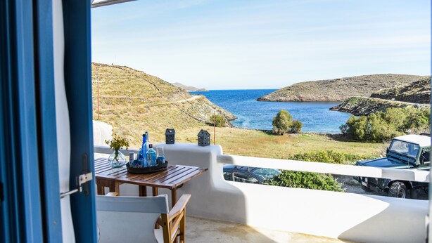 Uma varanda com vista para o mar, uma mesa com um vaso e garrafas, e um carro estacionado do lado de fora.
