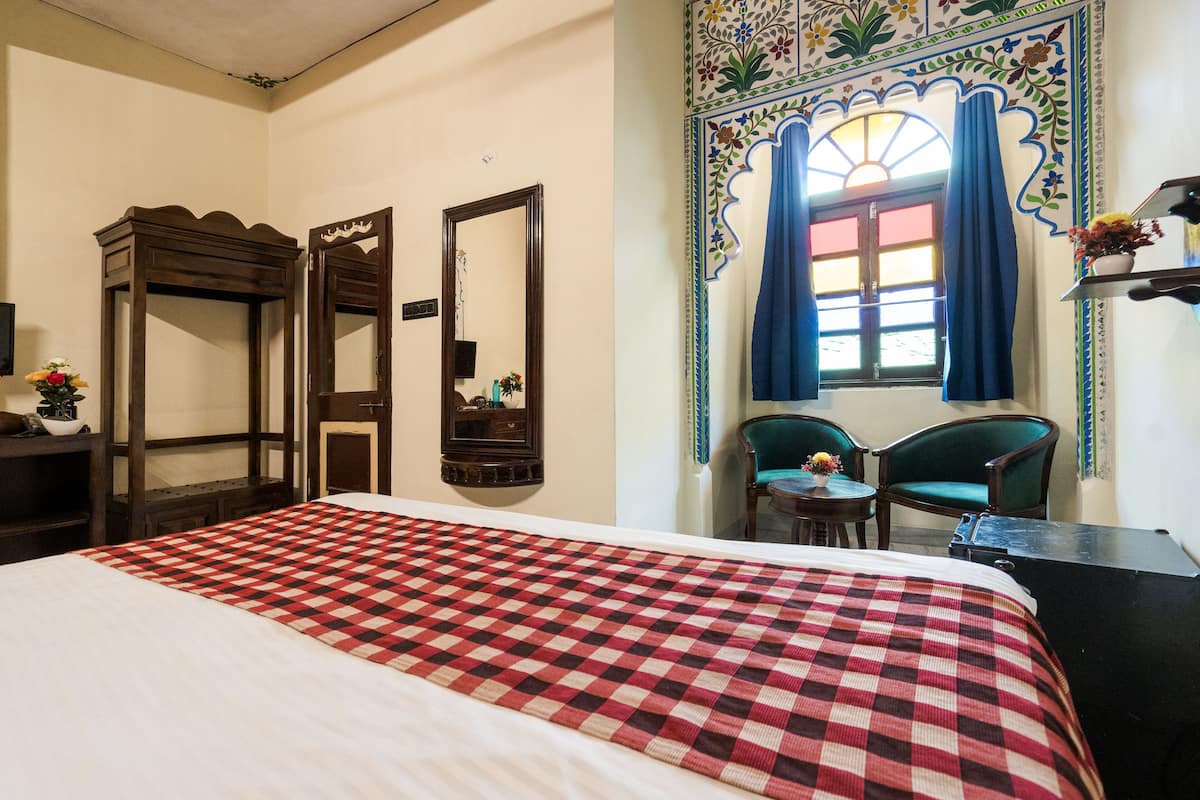 A hotel room with a checkered bedspread, two chairs, a desk, and a decorative mirror.
