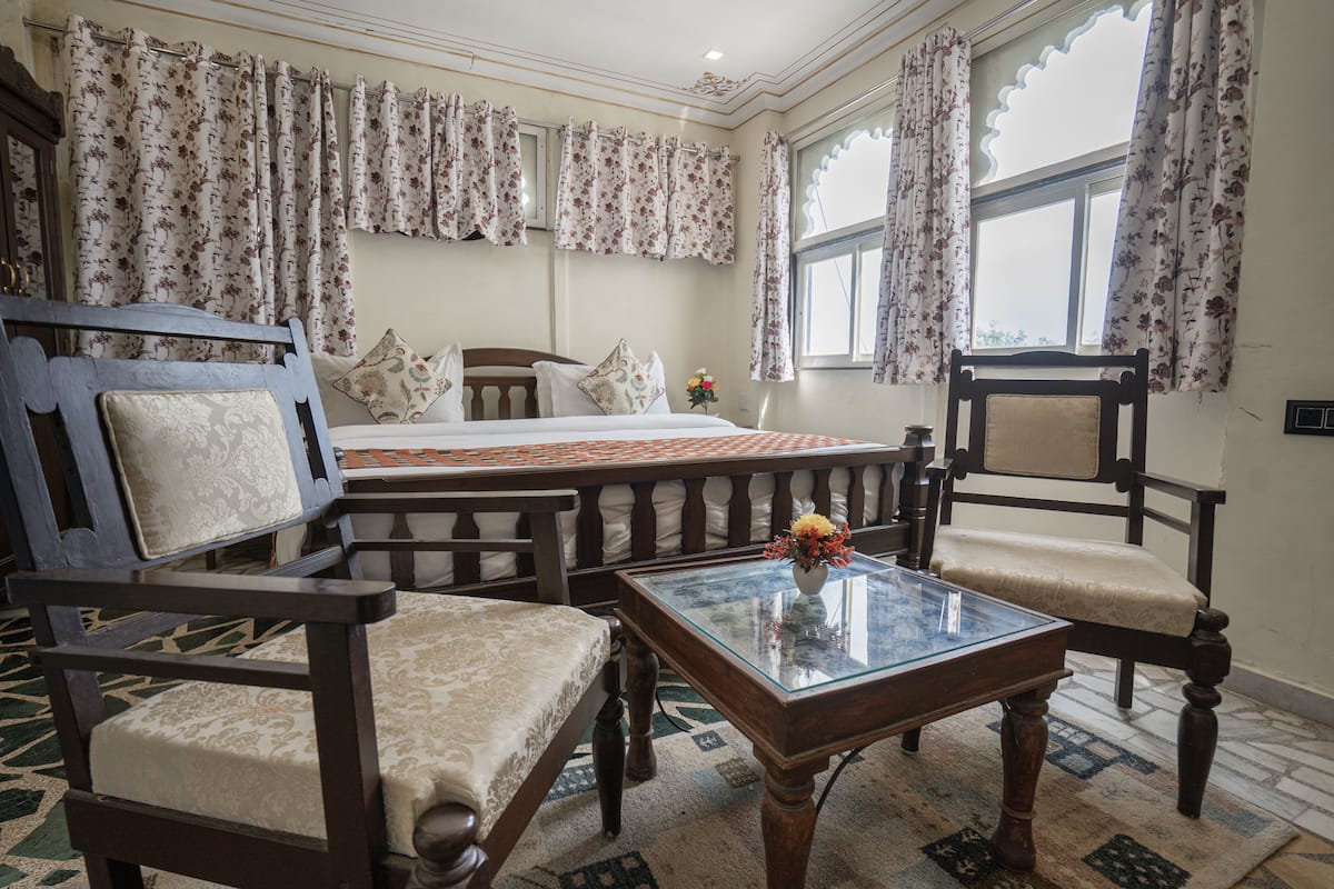 A hotel room with a bed, two wooden chairs, a glass coffee table, and floral curtains.