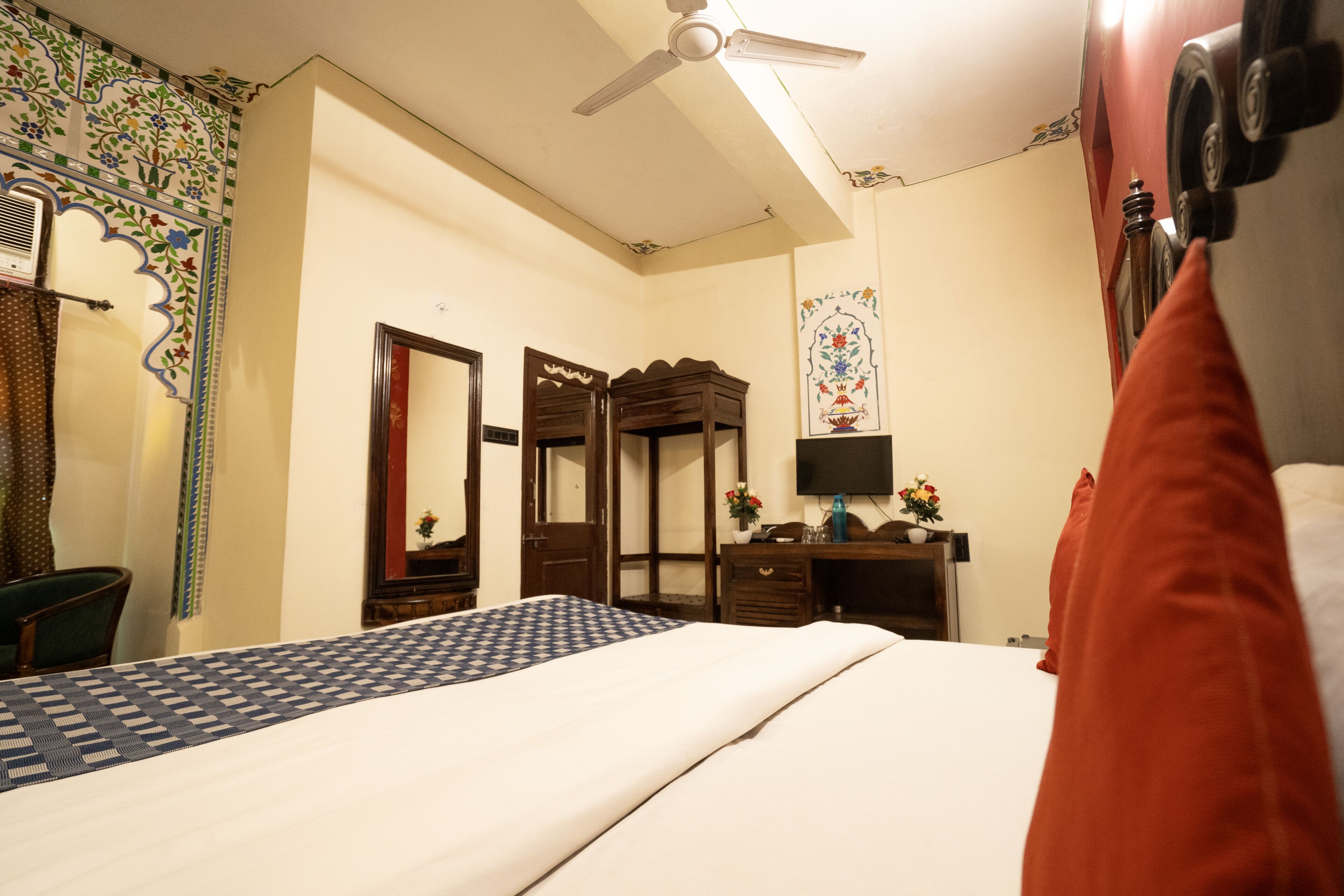 A hotel room with a bed, a desk, a chair, a wardrobe, a mirror, and a ceiling fan.