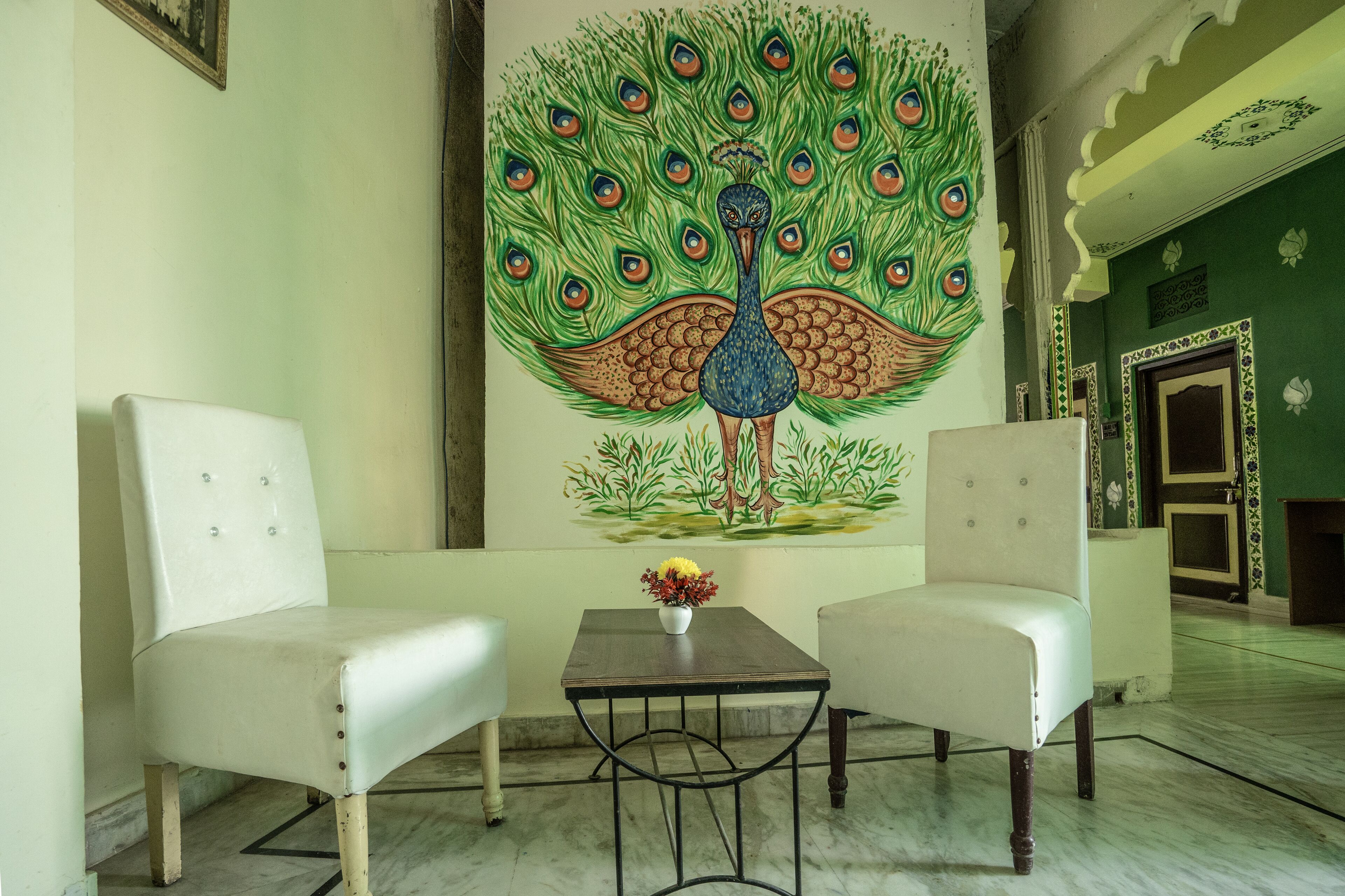 Una habitación con dos sillones blancos, una mesita con un jarrón de flores y un amplio mural de un pavo real en la pared.