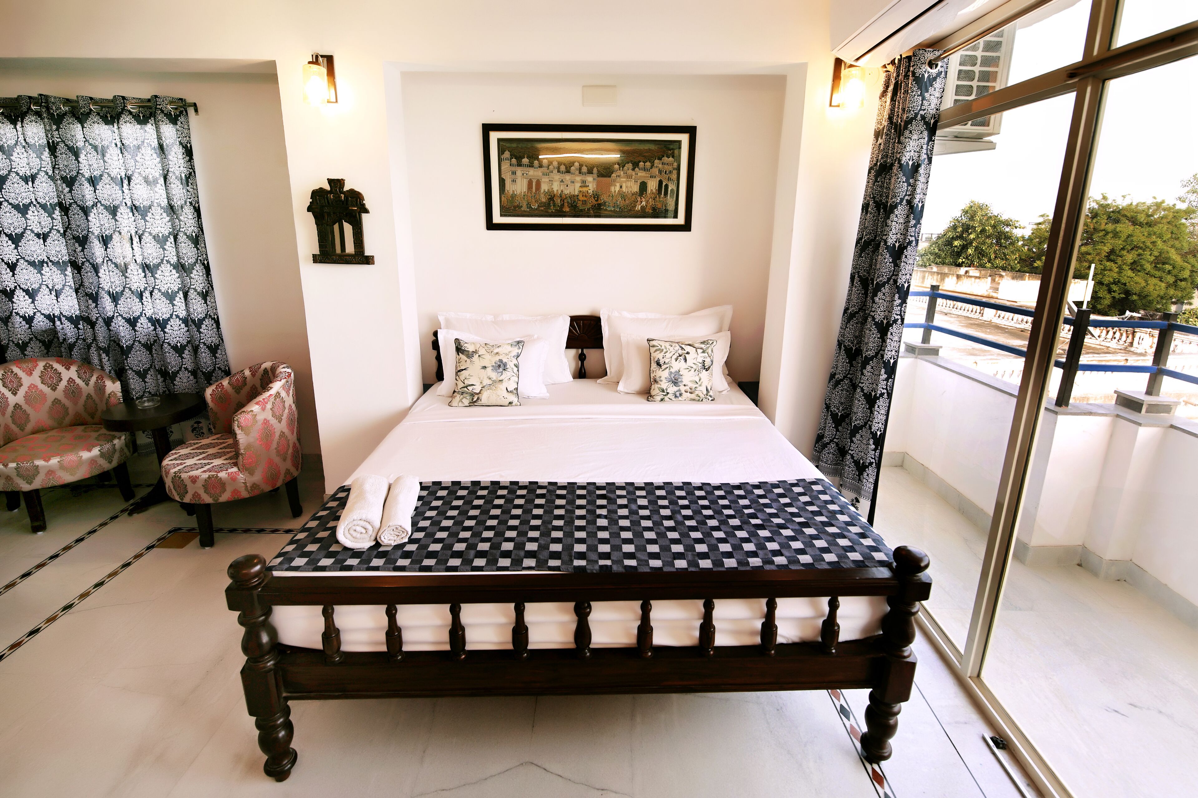 Un lit bien fait, recouvert d’une courtepointe à carreaux, une paire de chaises à motifs floraux, un tableau encadré au mur et un balcon avec une balustrade.