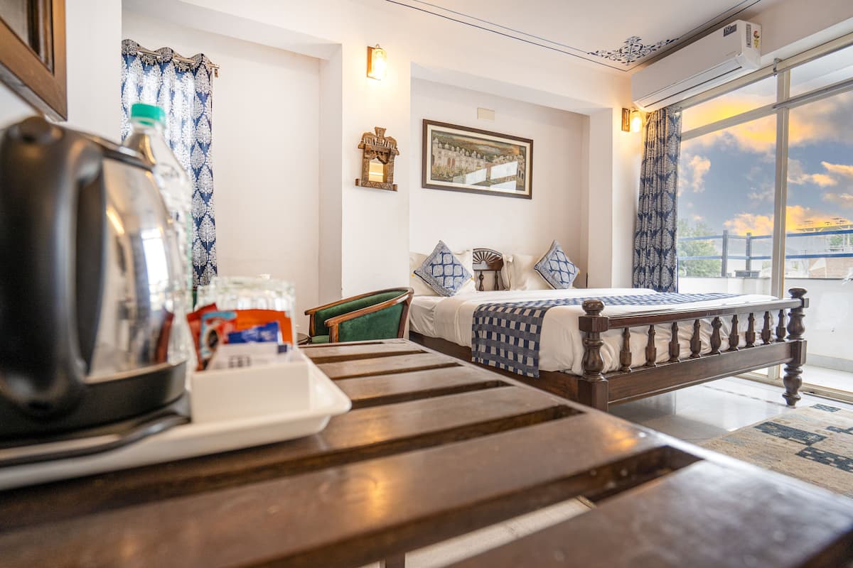 A hotel room with a wooden bed, a table with a kettle and a bottle, a window with a view, and a framed picture on the wall.