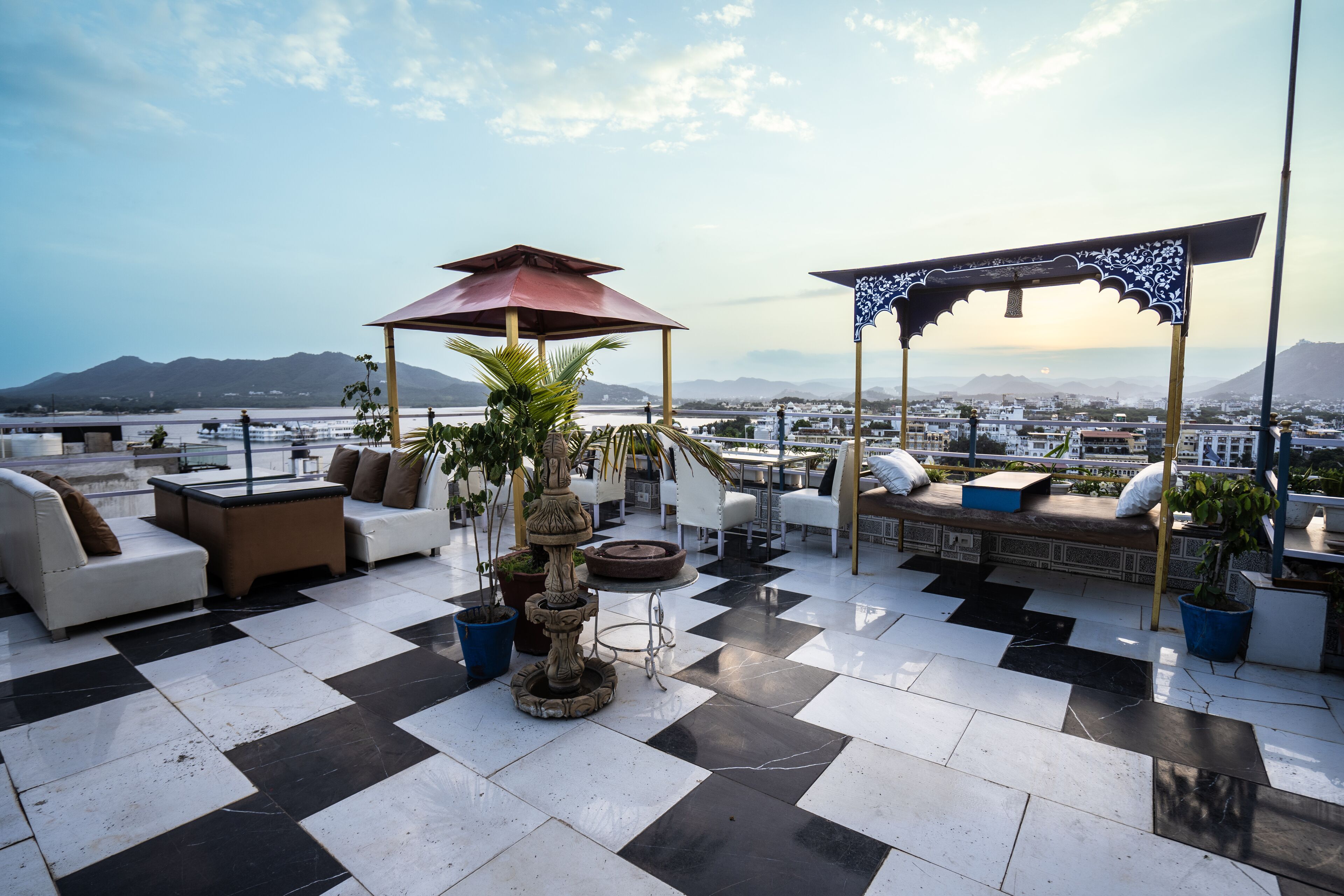 A rooftop terrace with checkered flooring, outdoor seating, a decorative fountain, and a view of a cityscape and mountains.