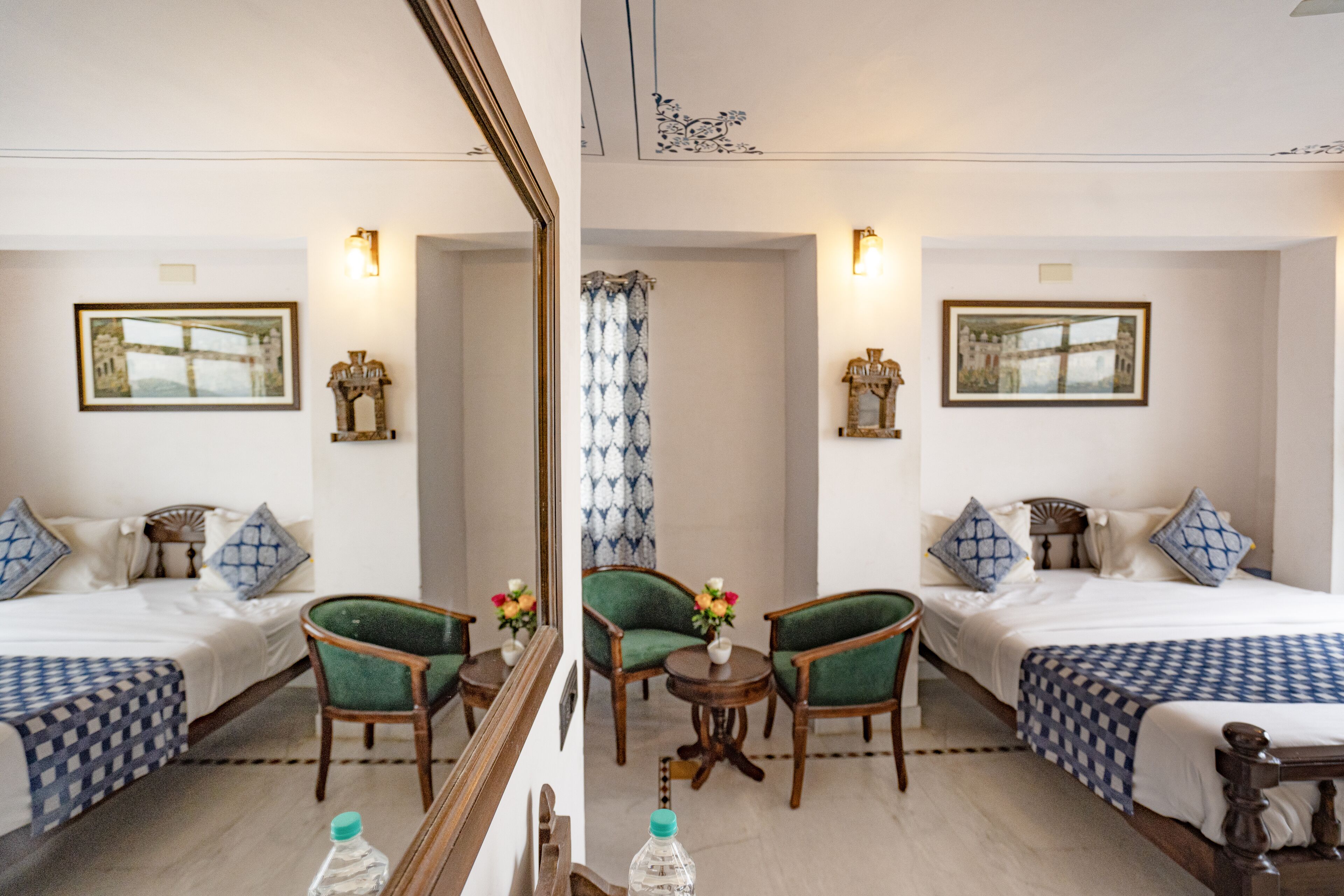 A hotel room with two beds, green upholstered chairs, a small table with a vase of flowers, and framed pictures on the walls.