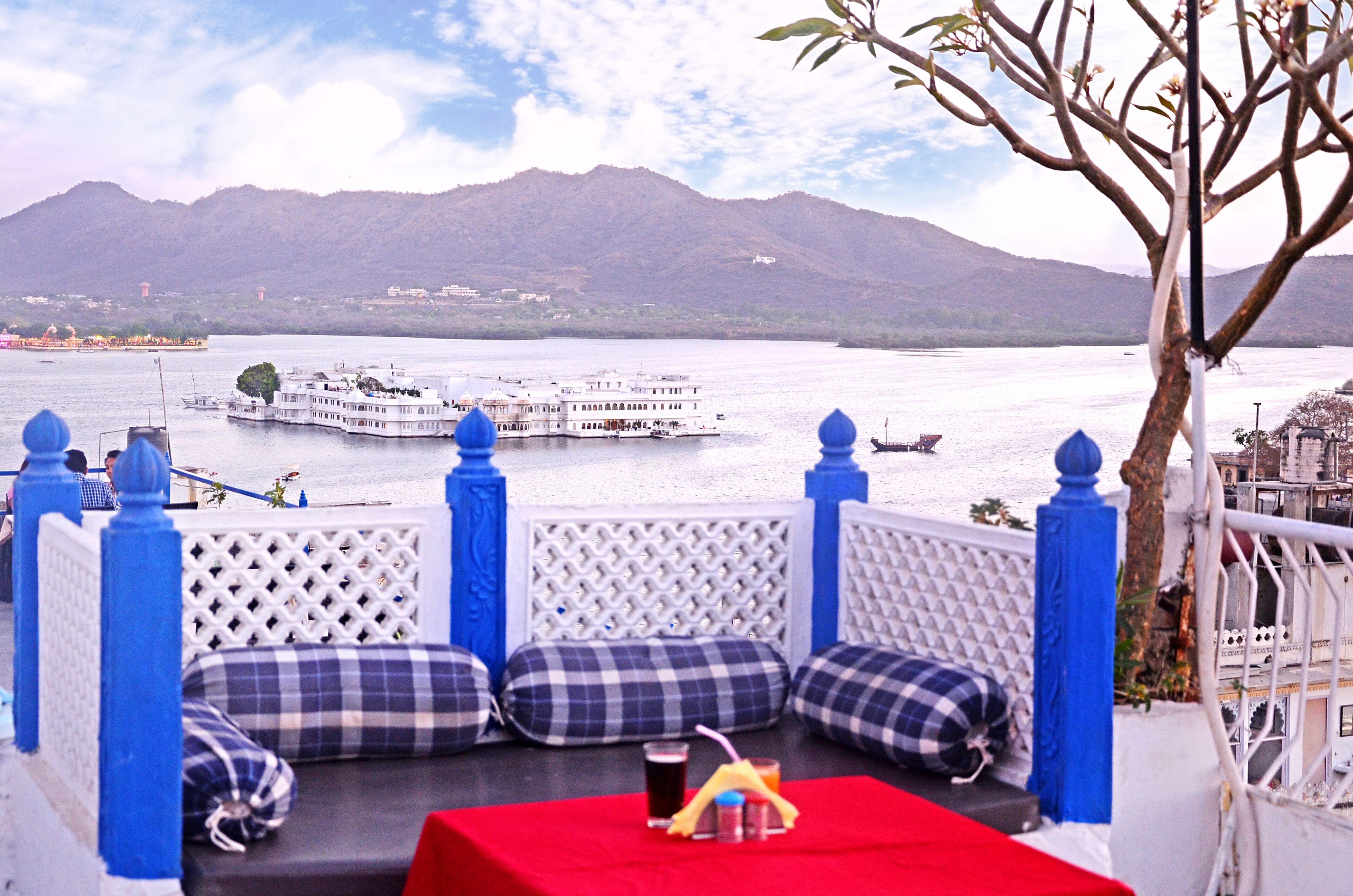 A terrace with a red table, checkered cushions, and a view of a lake with a boat and mountains in the background.