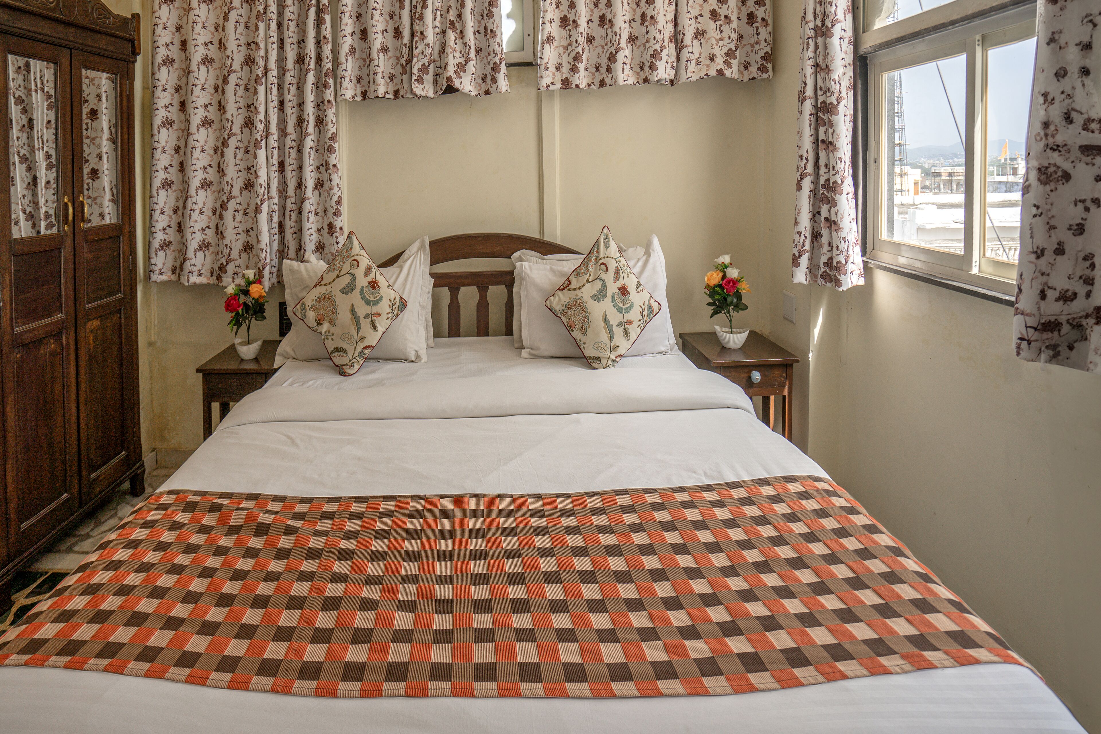 A neatly made bed with a checkered bedspread, floral curtains, and a view of the city through the window.