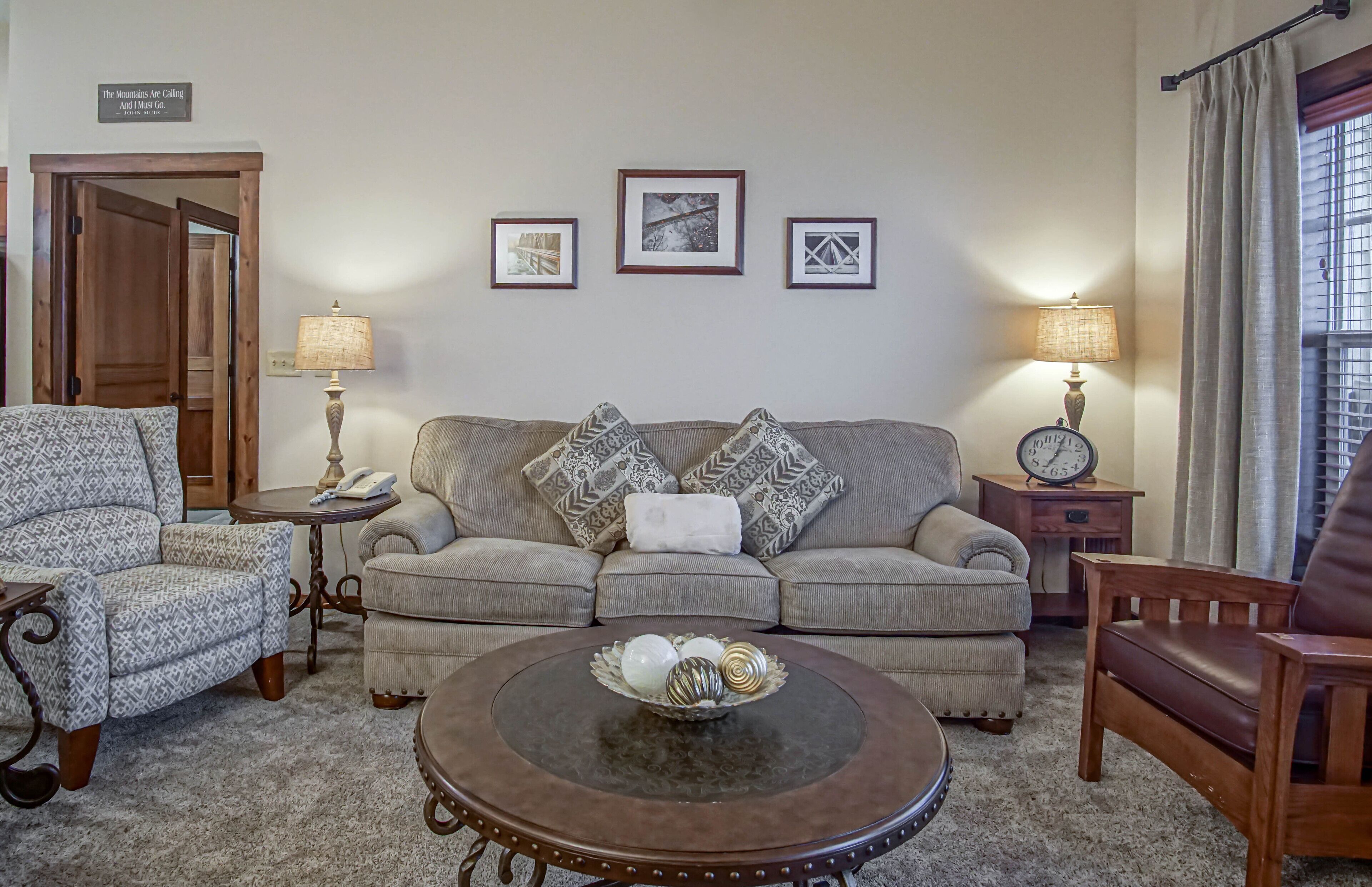 A living room with a patterned sofa, a round coffee table with decorative items, a patterned armchair, and a wooden side table with a lamp.
