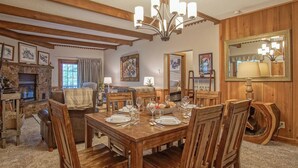Een eetkamer met een houten tafel voor zes personen, omringd door houten stoelen. Er is een stenen open haard met een schouw, en een kroonluchter hangt aan het plafond.