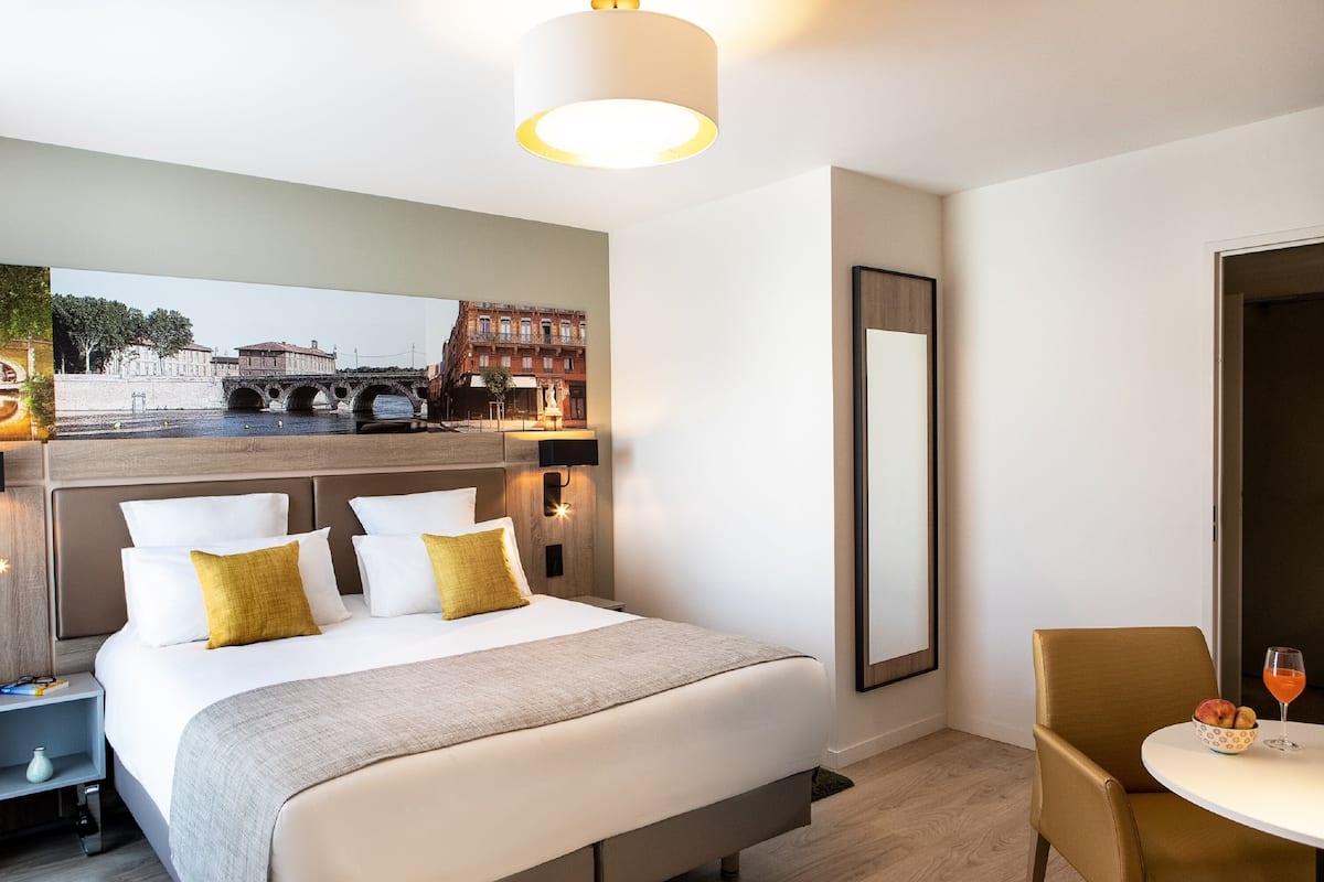 Ein Hotelzimmer mit einem großen Bett, einem Nachttisch, einem Stuhl, einem kleinen Tisch mit einer Obstschale und einem Glas Orangensaft sowie einem großen Spiegel.