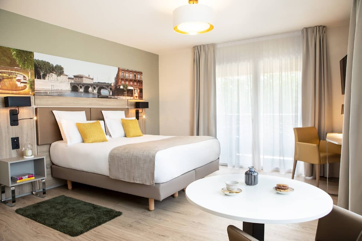 Ein modernes Hotelzimmer mit einem großen Bett, einem runden Esstisch und Blick auf eine Brücke sowie auf Gebäude durch das Fenster.