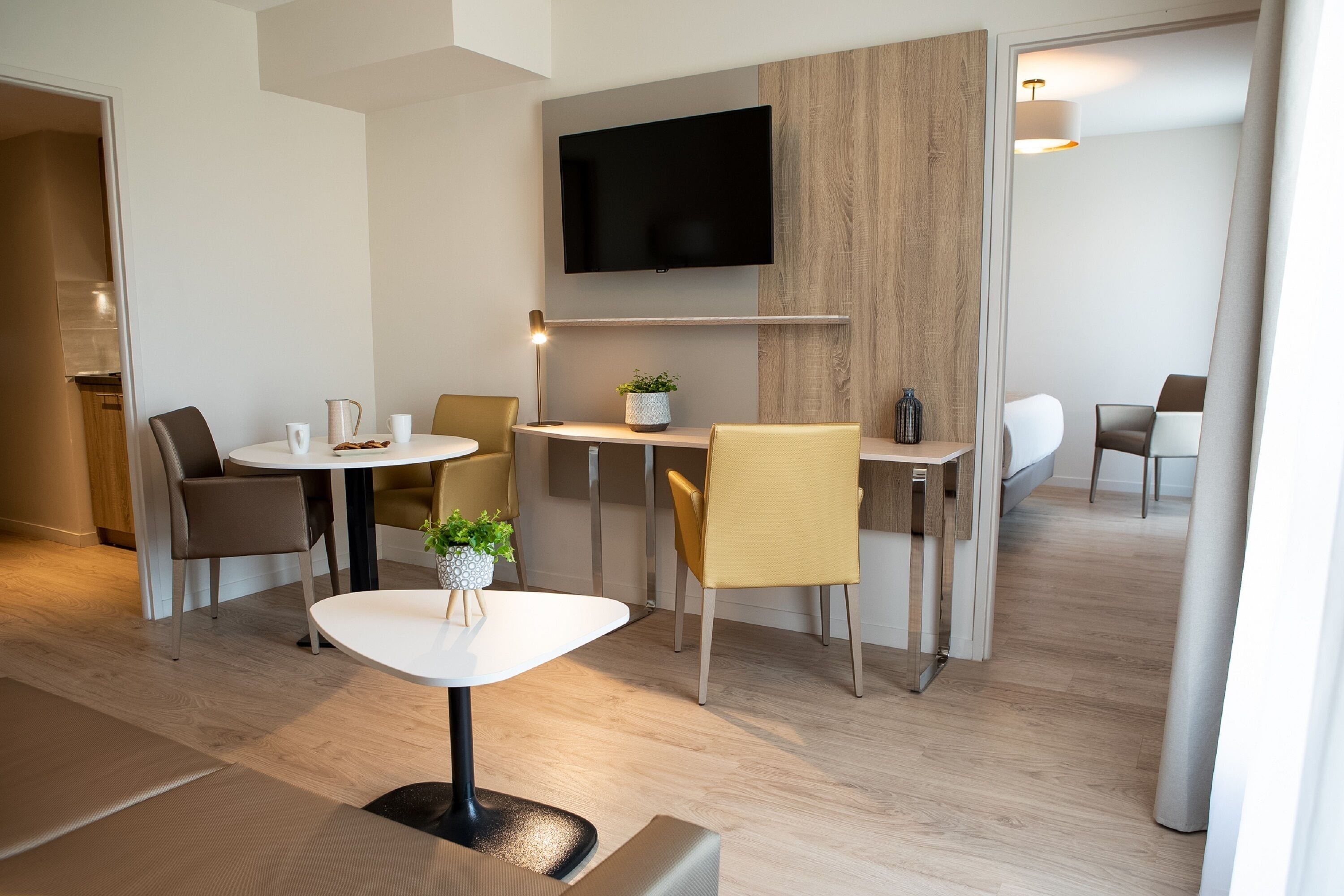 A modern hotel room with a flat-screen TV mounted on the wall, a round dining table with chairs, and a sofa.