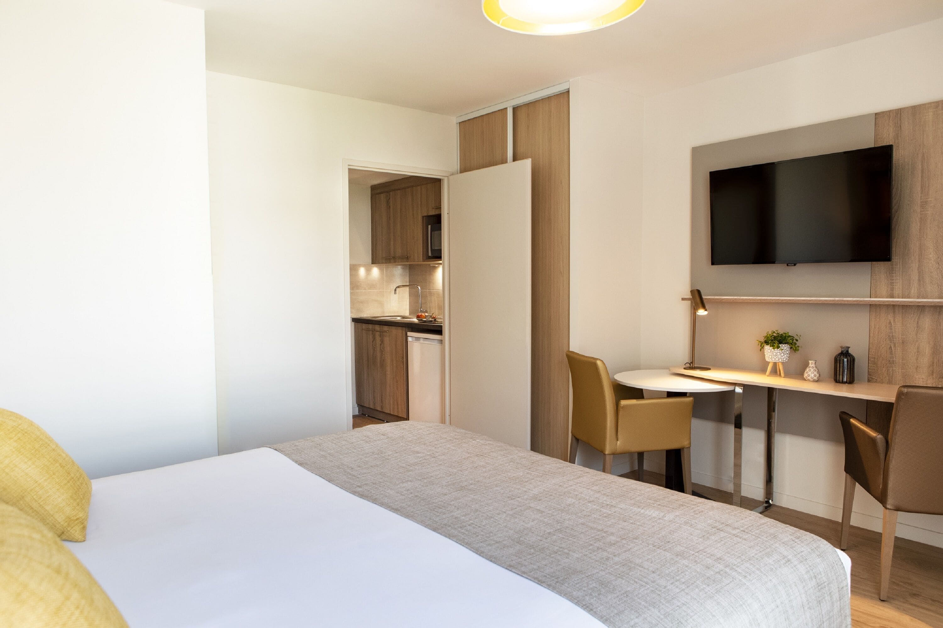 A modern hotel room with a bed, a desk, a chair, a television, and a kitchenette in the background.