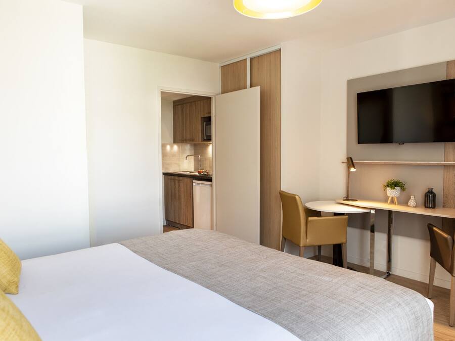 A modern hotel room with a bed, a desk, a chair, a television, and a kitchenette in the background.