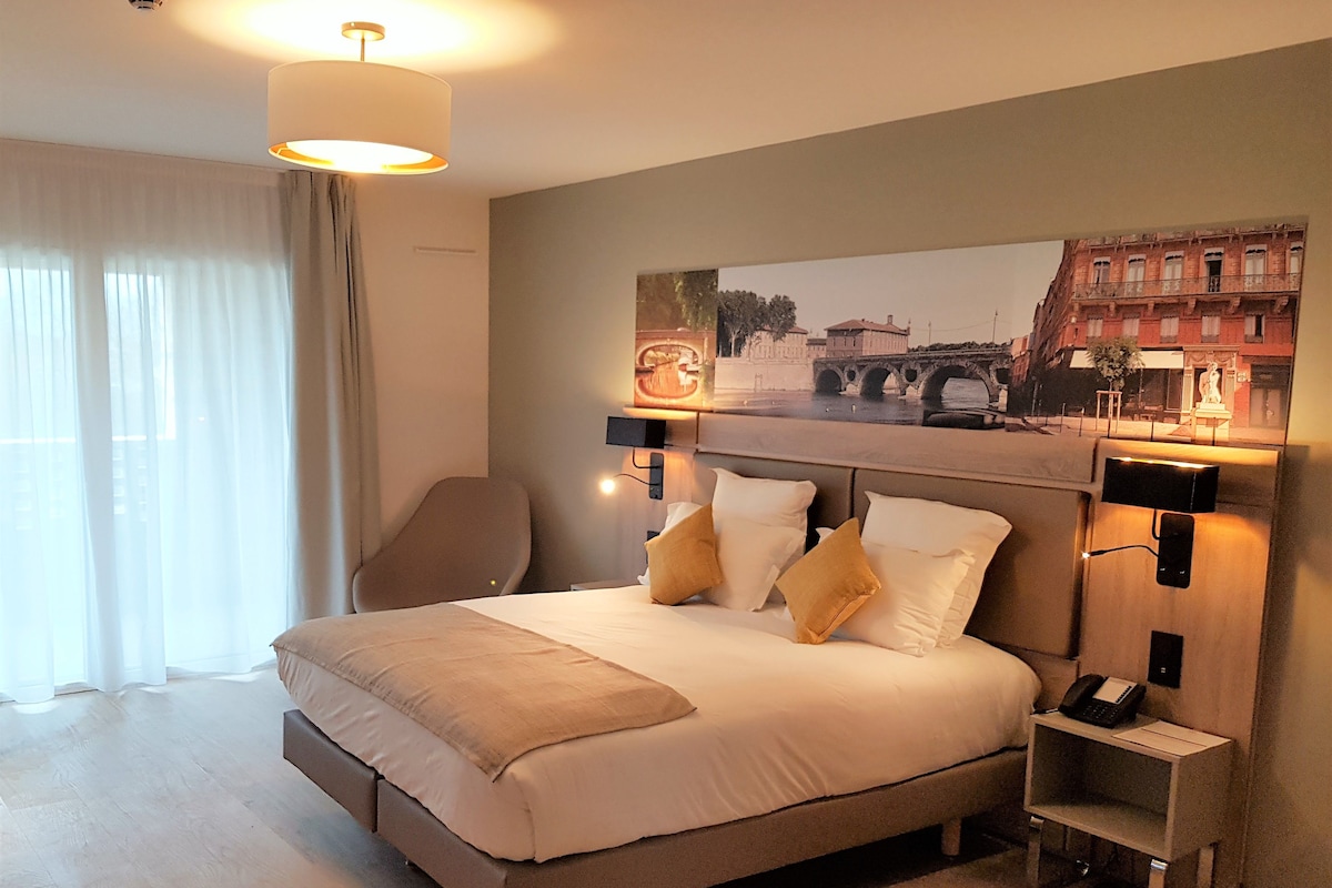 A hotel room with a large bed, a desk with a phone, and a painting of a bridge on the wall.