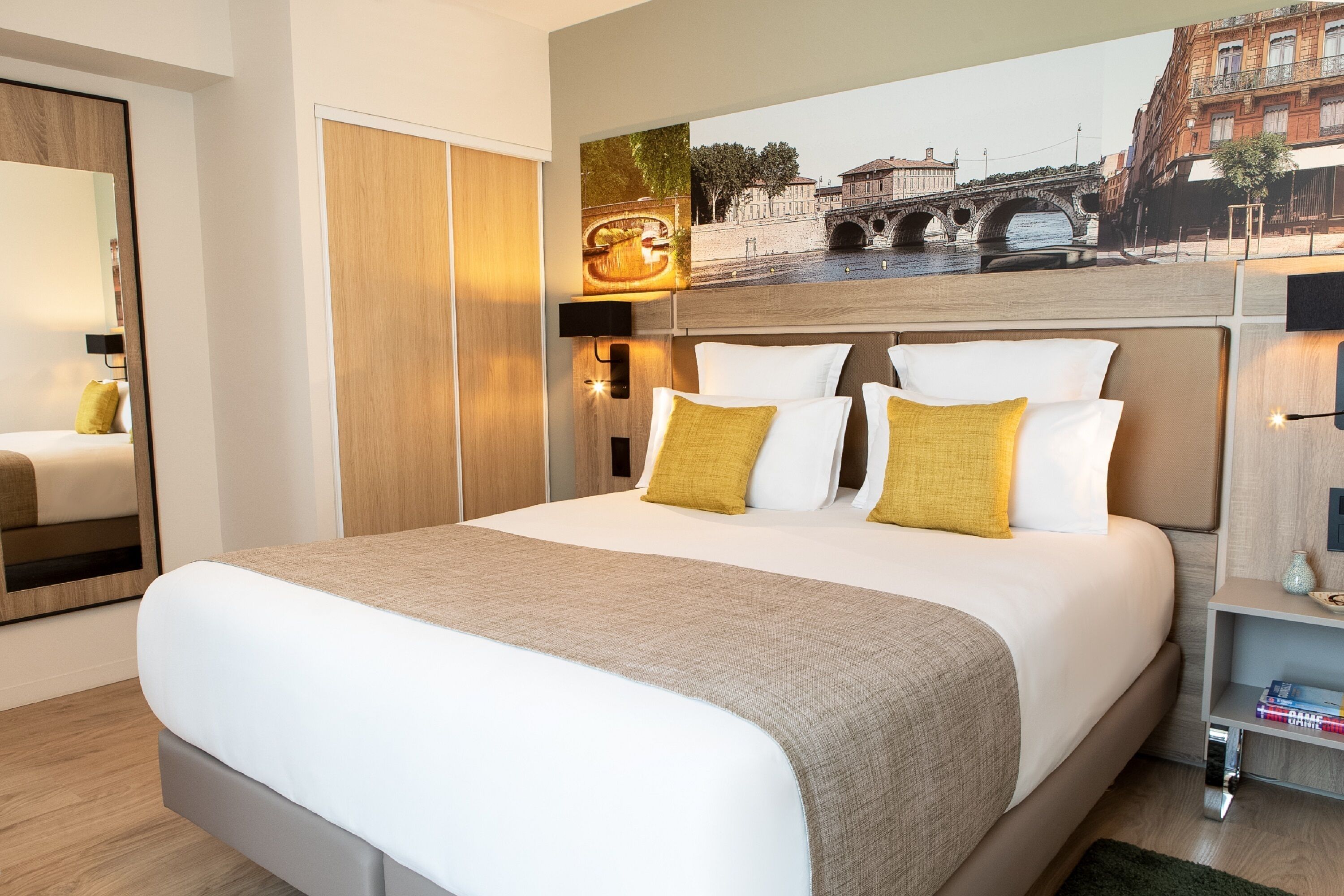 Une chambre d’hôtel moderne avec un grand lit, une tête de lit en bois et une fresque murale représentant un paysage urbain.