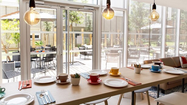 A dining area with a long table set for four, large windows, and hanging light fixtures. - Residhome Toulouse Ponts Jumeaux (Toulouse)