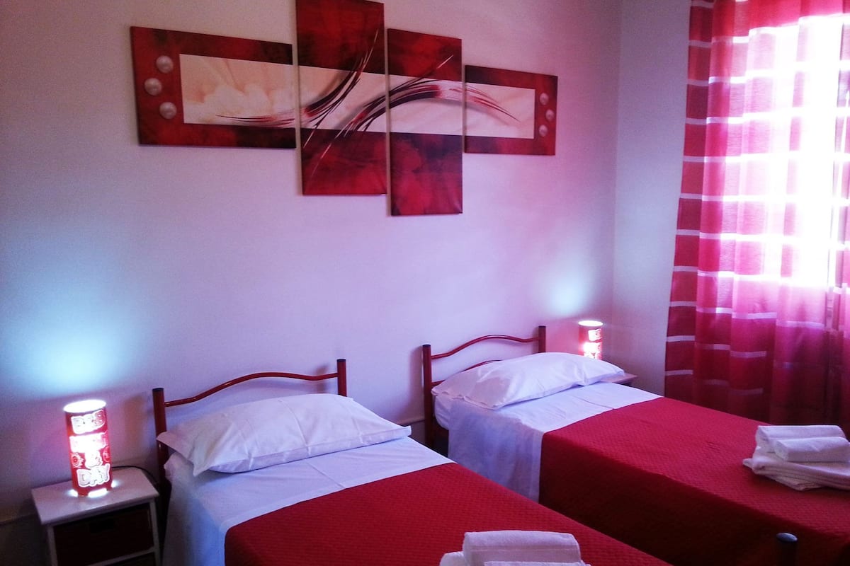 A hotel room with two beds, each with a red bedspread, white pillows, and a nightstand with a lamp. There is a wall art piece above the beds and a window with red curtains.