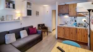 Un espacio habitable compacto con sofá, una pequeña mesa de comedor, una kitchenette con armarios de madera y una estantería.