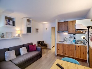 A compact living space with a sofa, a small dining table, a kitchenette with wooden cabinets, and a bookshelf.