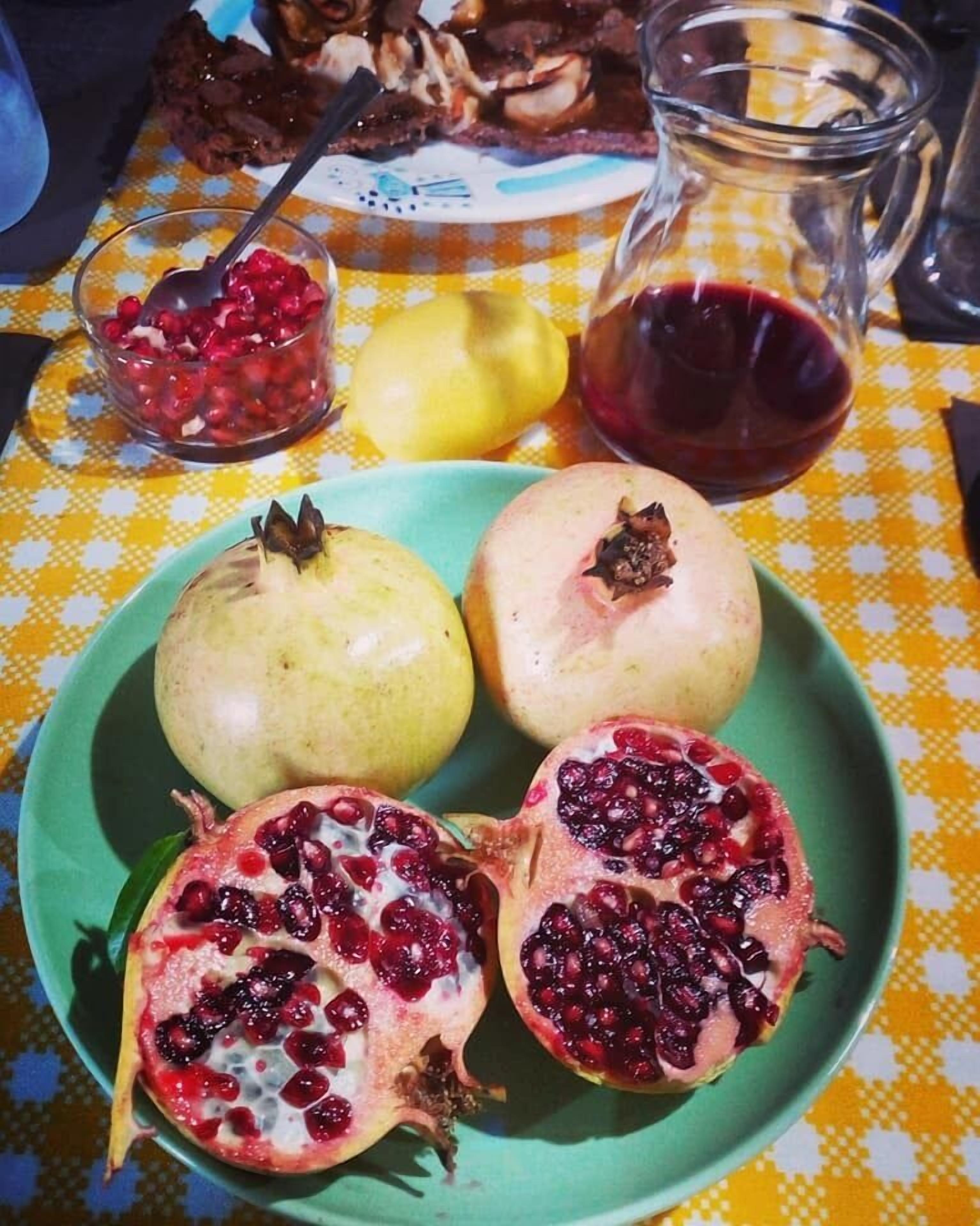 A table with a checkered tablecloth, a green plate with pomegranates, a lemon, and a glass pitcher.