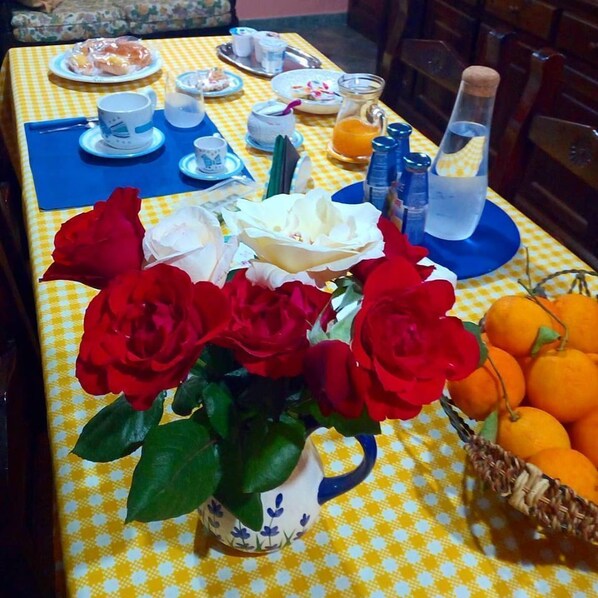 Una mesa vestida con mantel a cuadros, un jarrón de rosas rojas y blancas, una cesta de naranjas y diferentes platos y bebidas.