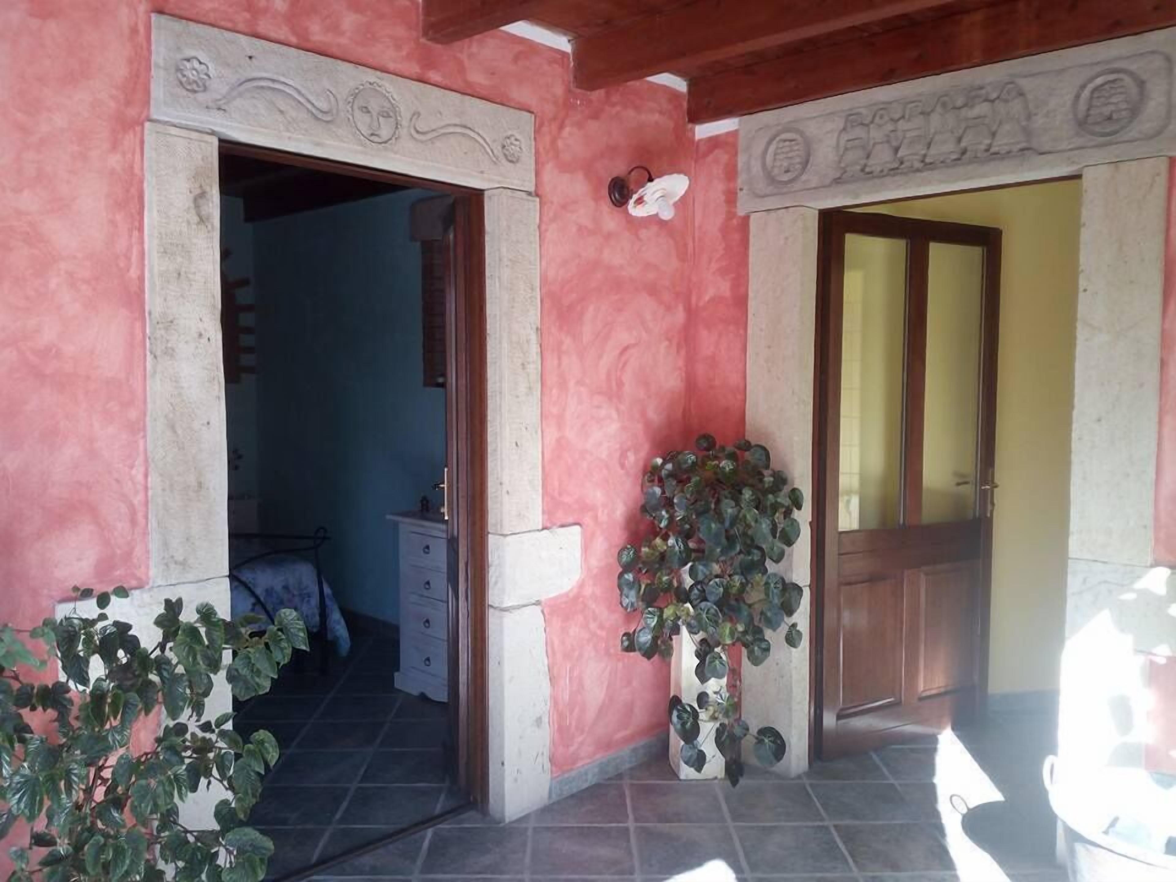 Una stanza con una parete rosa, una porta in legno e un pavimento piastrellato. Nell'angolo c'è una pianta e un piccolo tavolo con cassetti.