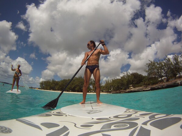 Two people paddleboarding on a clear blue water body with a tropical landscape in the background.