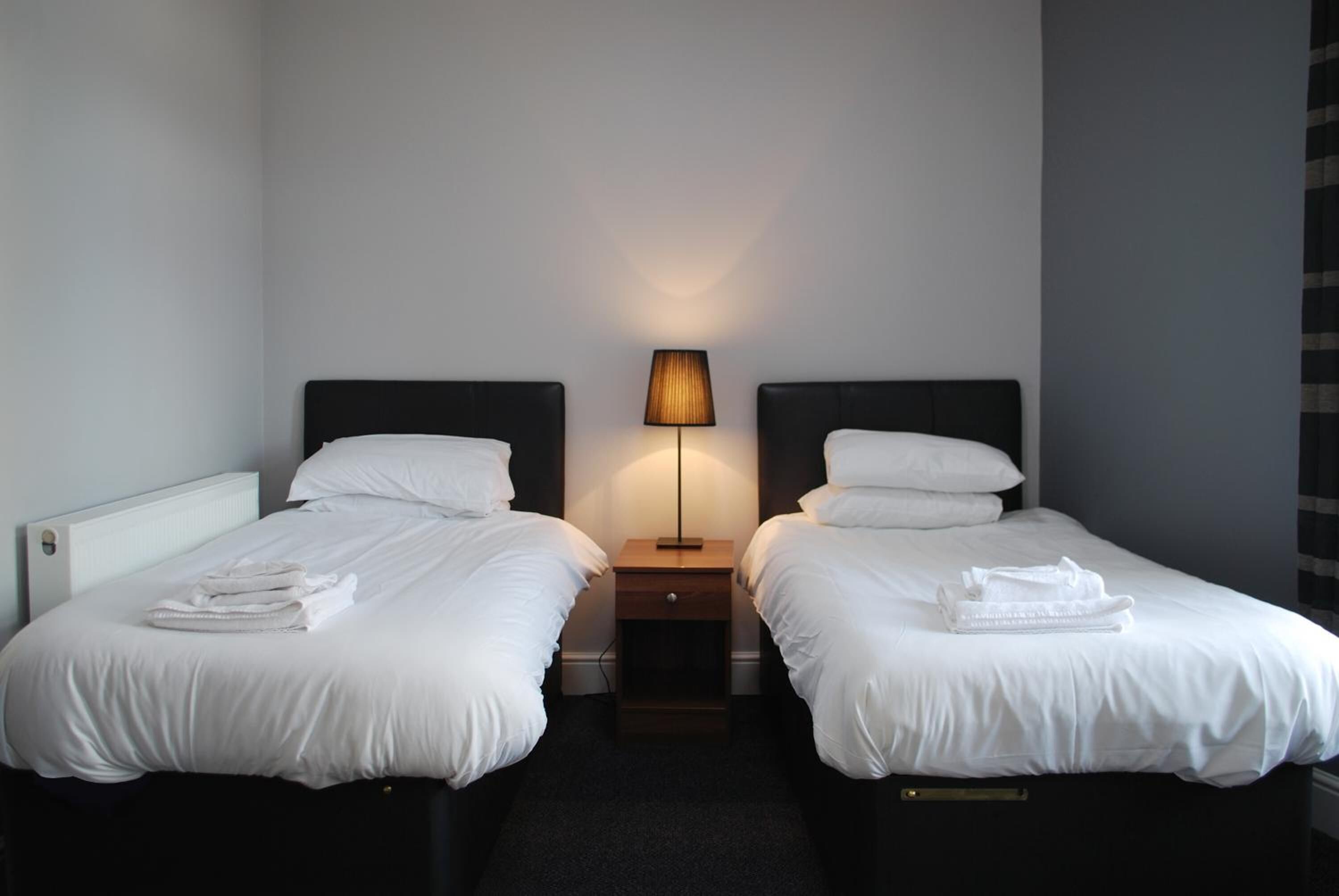 A hotel room with two beds, a nightstand with a lamp, and a radiator.