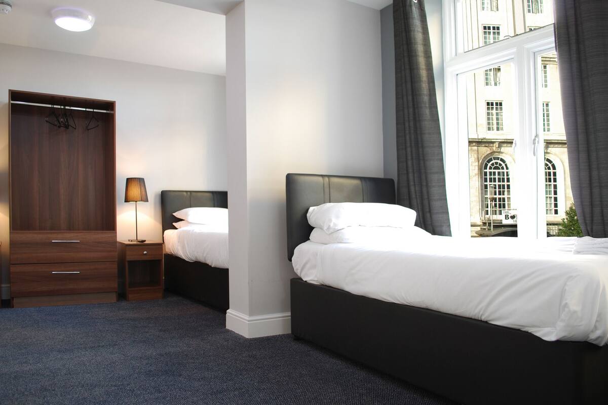 A hotel room with two beds, a wardrobe, a nightstand with a lamp, and a view of a building through a window.