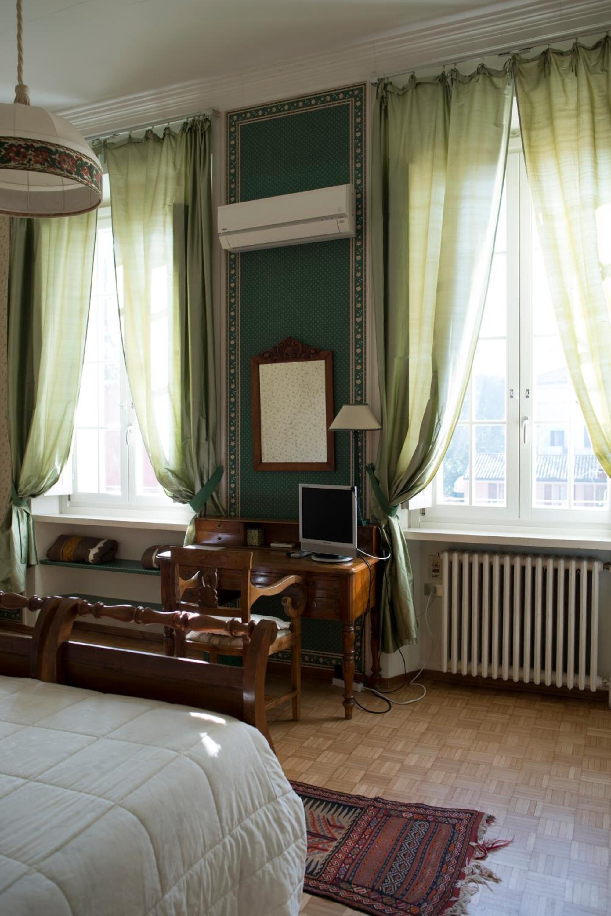 A room with a bed, a desk, a chair, a radiator, and a window with curtains.