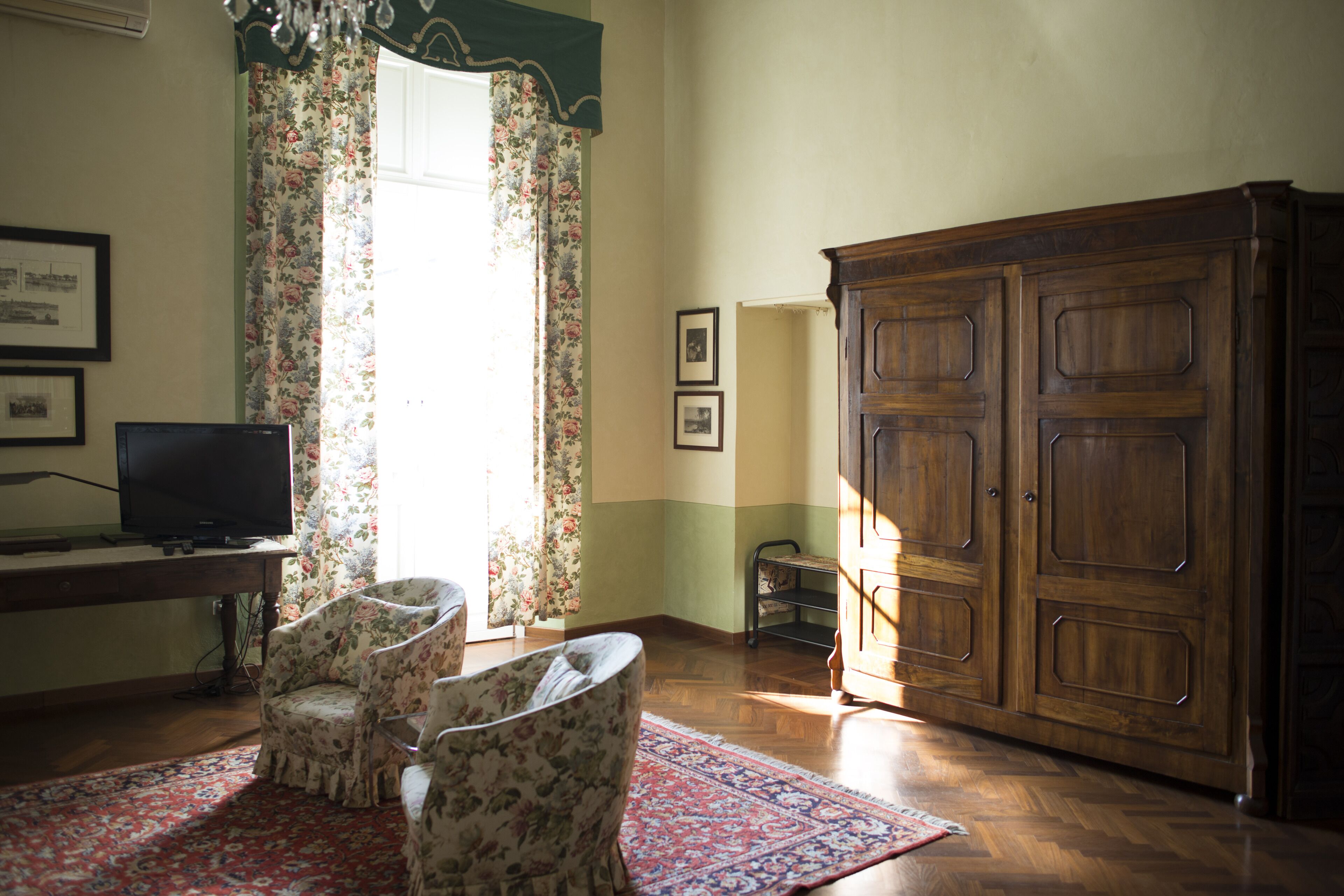 A room with a wooden cabinet, two floral armchairs, a television on a desk, and a patterned rug.