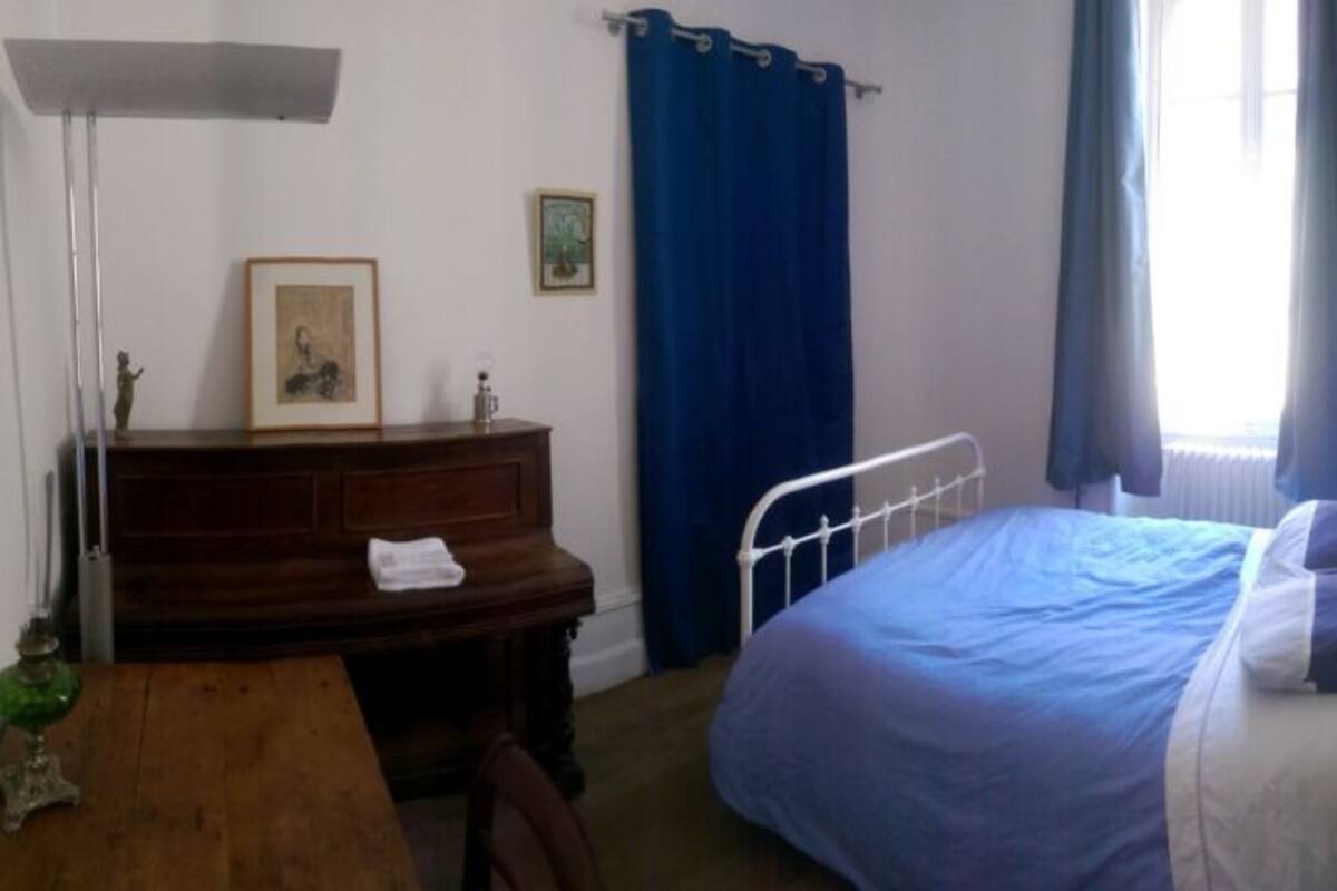 Ein Schlafzimmer mit einem Bett, einer Holzkommode, einem Schreibtisch, einer Lampe und einem Fenster mit Vorhängen.