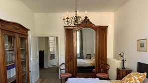 A bedroom with a large bed, a wooden wardrobe, a mirror, a chandelier, and a nightstand with a lamp. - Grand Corps de Ferme (Branches)