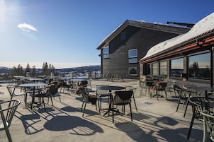 Um pátio externo com mesas e cadeiras, um edifício com grandes janelas, e vista para uma montanha nevada.