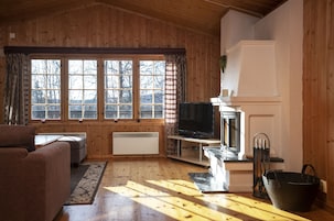 Interior acolhedor de uma cabana, com decoração em madeira, lareira, televisão e uma grande janela com cortinas.