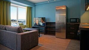 A modern living room with a sofa, a television, a kitchen area with a refrigerator and microwave, and a view of a cityscape through large windows. - Skeikampen Servicesenter (Gausdal)
