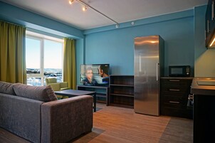 Uma sala de estar moderna com sofá, televisão, área de cozinha com frigorífico e micro-ondas, e vista para a paisagem urbana através de amplas janelas.