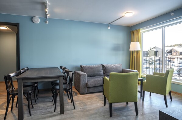 A room with a dining table, chairs, a sofa, and a window with a view of parked cars. - Skeikampen Servicesenter (Gausdal)