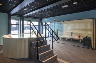 Um hall moderno com um balcão de recepção curvo, uma escadaria com guarda-corrimão de metal, uma área de estar com um banco e um painel de vidro.