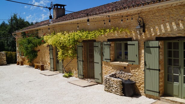A rustic stone building with a tiled roof, green shutters, and a gravel courtyard.