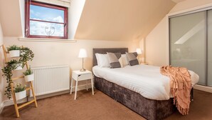 Ein Schlafzimmer mit Bett, Nachttischlampen, Heizkörper, einem Fenster mit Blick und einem Regal mit Pflanzen.
