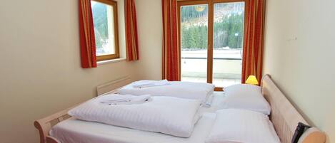 A double bed with white linens, two windows with red curtains, a wooden headboard, and a bedside table with a lamp.