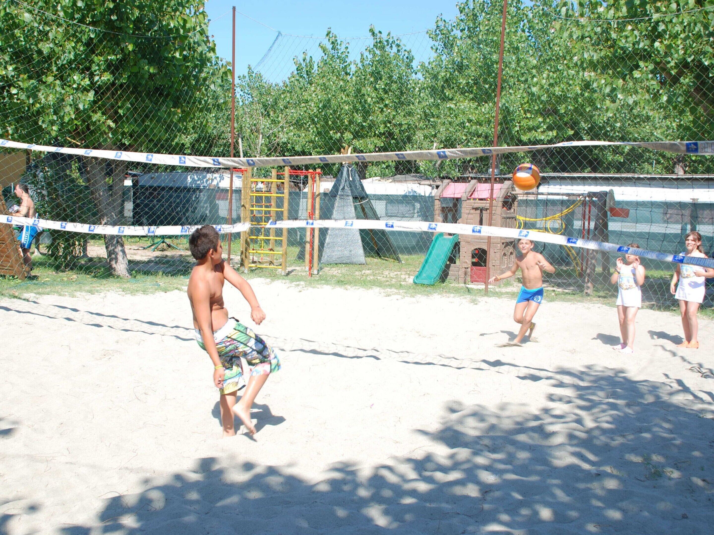 Un match de volleyball est en cours sur un terrain de sable entouré d’une clôture en treillis métallique.